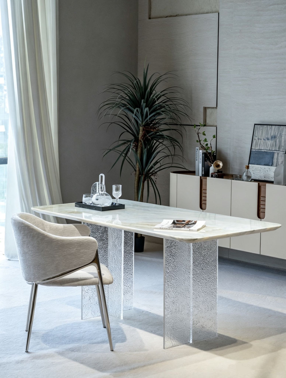 A modern dining table with a white marble top and textured clear glass legs, paired with a light beige upholstered chair in a minimalist, contemporary dining room setting.