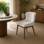 A modern upholstered dining chair with a white curved seat and back, featuring vertical channel tufting and tapered brown wooden legs, set in a warm, sunlit dining room next to a round wooden table.