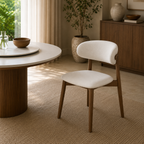 A modern dining chair with a curved white upholstered seat and backrest, supported by tapered medium-brown wooden legs, shown in a styled dining room next to a round white table.