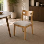 A light natural wood dining chair with a square off-white upholstered seat, shown in a bright, modern dining room setting next to a round table.