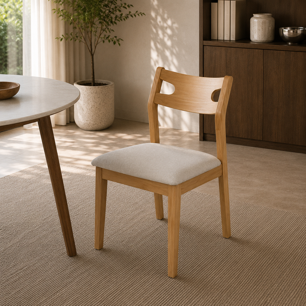 A light natural wood dining chair with a square off-white upholstered seat, shown in a bright, modern dining room setting next to a round table.