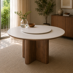 A round dining table with a light beige tabletop and thick, sculptural brown wooden legs, styled in a warm, modern dining room with a matching round tray on top.