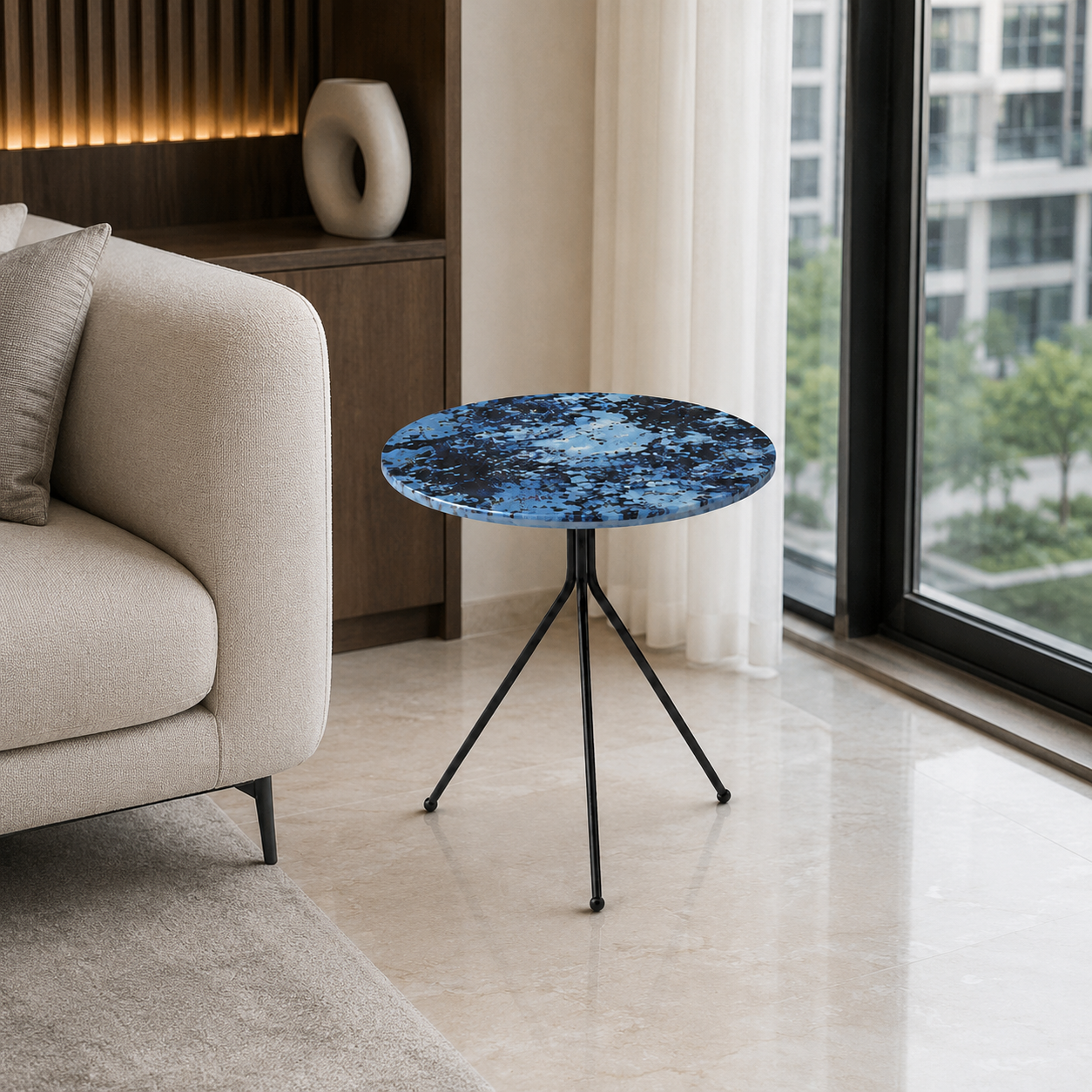 A small round side table with a blue marbled top and black tripod legs, placed in a modern living room next to a beige sofa and a large window.