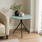 A round side table with a mint green top and black metal tripod legs, styled in a living room corner next to a beige armchair with a potted plant and a small cup on top.