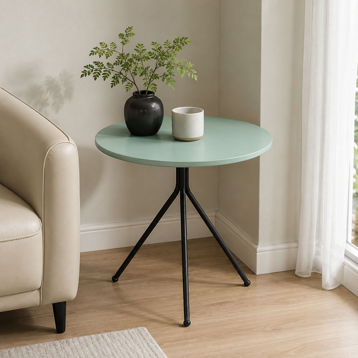 A round side table with a mint green top and black metal tripod legs, styled in a living room corner next to a beige armchair with a potted plant and a small cup on top.