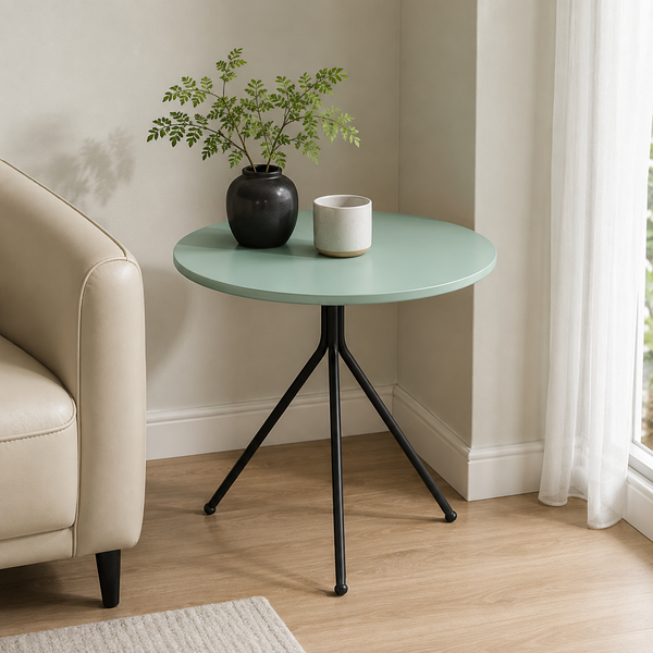 A round side table with a mint green top and black metal tripod legs, styled in a living room corner next to a beige armchair with a potted plant and a small cup on top.