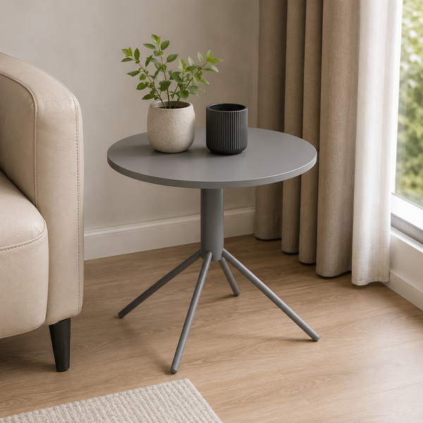 A round gray side table with a three-legged metal tripod base, styled in a living room next to a beige armchair with a potted plant and a dark gray cup on top.