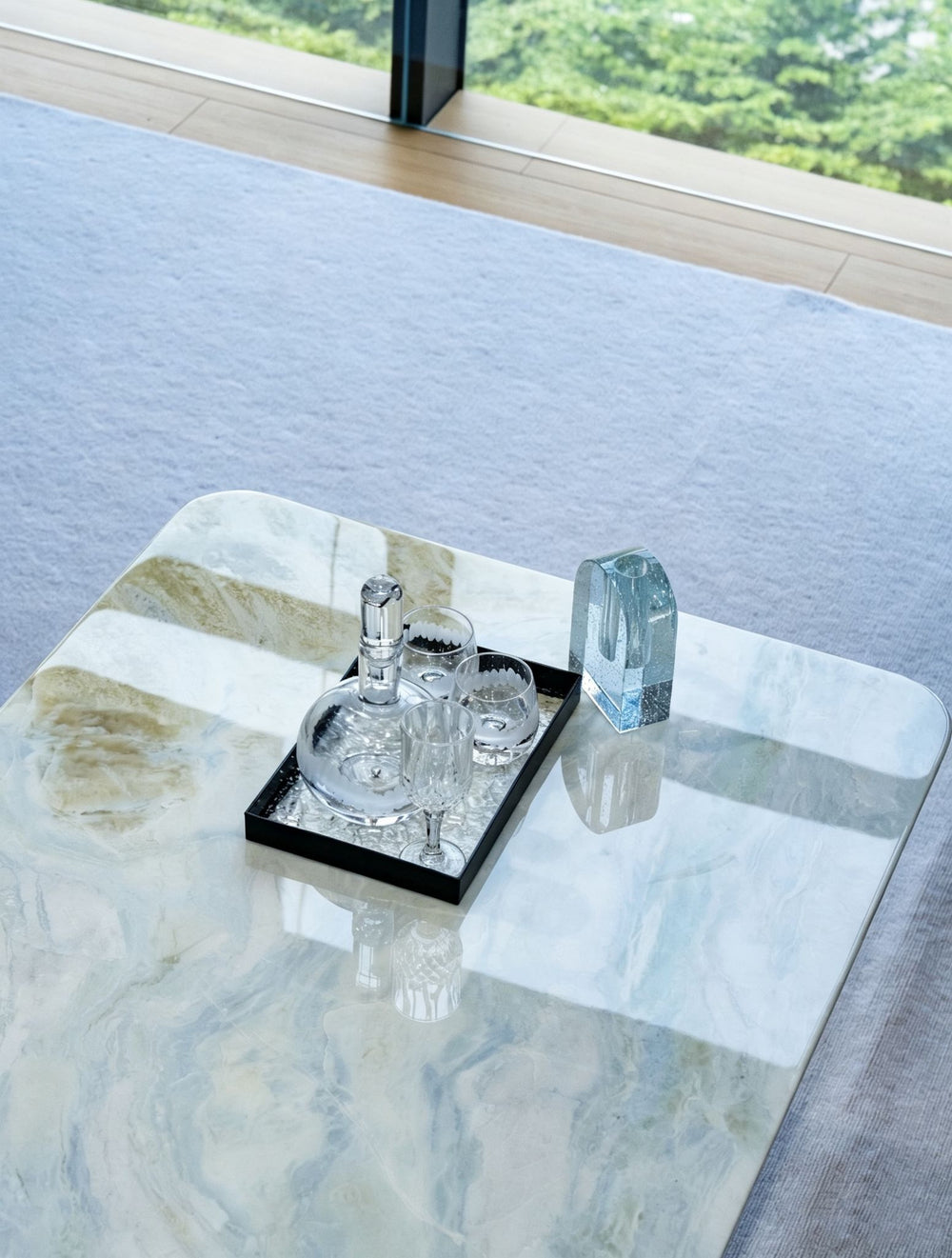 A modern rectangular coffee table with a white and gray marbled stone top, styled with a glass decanter, two small glasses on a black tray, and a clear glass vase, set in a bright room with a large window.