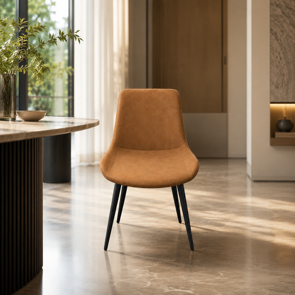 A modern accent chair with a warm camel-brown suede or velvet upholstered seat and back, supported by slim black metal legs, set in a stylishly decorated interior room with a marble floor and a curved wooden table.