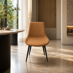 A modern accent chair with a warm camel-brown suede or velvet upholstered seat and back, supported by slim black metal legs, set in a stylishly decorated interior room with a marble floor and a curved wooden table.