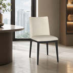 A modern dining chair with a cream upholstered seat and backrest and dark charcoal legs, positioned in a contemporary room with a large window showing a city skyline.