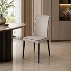 A modern dining chair with light beige upholstered seat and backrest and black legs, shown in a contemporary dining room setting next to a marble-top table.