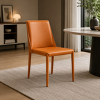 A modern side chair upholstered in warm orange-brown leather with matching tapered wooden legs, shown in a styled dining room setting next to a round table on a light rug.