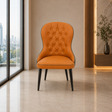 An orange tufted upholstered accent chair with black legs, positioned in a modern interior setting with a city view window on the left and a wooden console on the right.