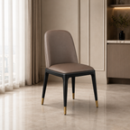 A modern dining chair with a taupe-brown upholstered seat and curved backrest, supported by black tapered legs with gold-tone metal caps, set in a contemporary interior room.