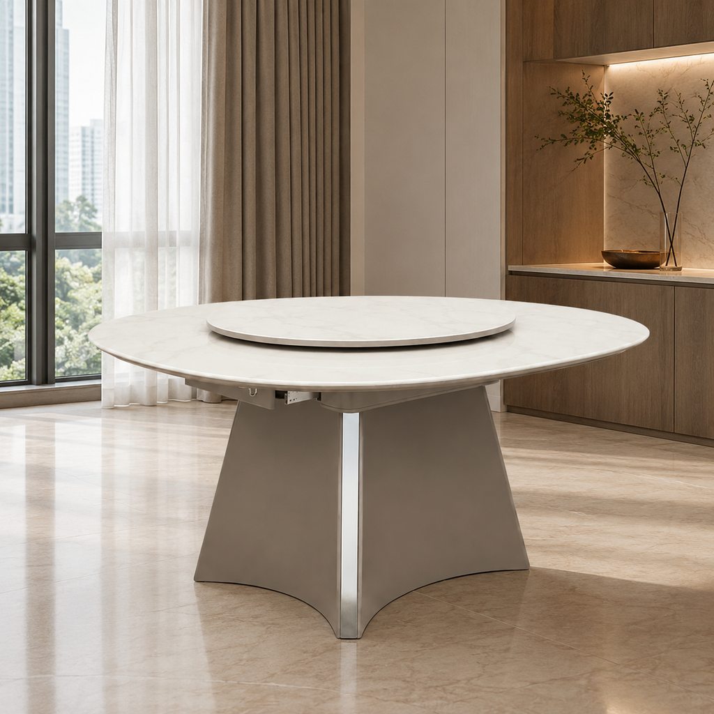 A modern round dining table with a light stone-look top and a sculptural metallic base, shown in a bright, contemporary interior with large windows and warm wood cabinetry.