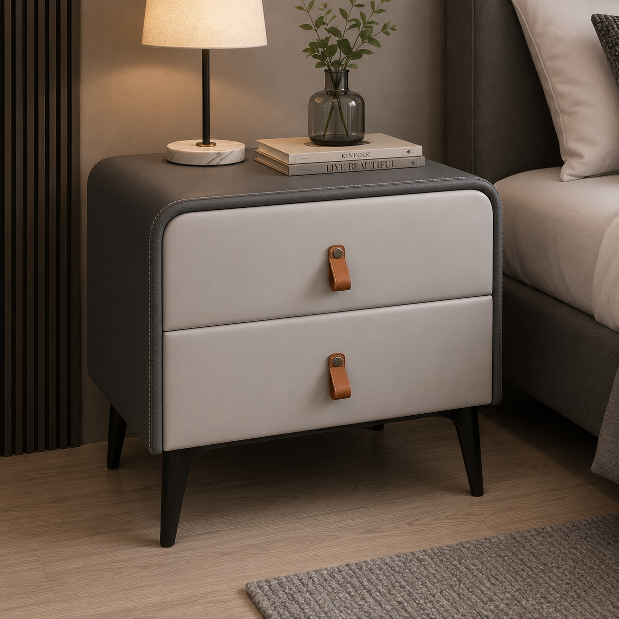 A modern two-drawer nightstand with a dark gray faux-leather body, light gray front panels, and brown leather pull handles, styled in a bedroom next to a bed with a lamp and books on top.