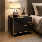 A modern black nightstand with gold trim and legs, styled in a bedroom next to a bed with white linens. The nightstand features a glossy finish, a lower open shelf, and two drawers with slim gold handles.
