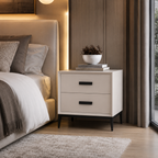 A modern two-drawer nightstand in a warm off-white finish with black handles and tapered black legs, styled in a contemporary bedroom next to a bed with neutral-toned bedding.
