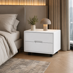A modern white two-drawer nightstand with black caster wheels, styled in a bedroom next to a bed with beige bedding, a small potted plant, and a dome-shaped table lamp.