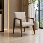 A modern upholstered accent chair with a light beige fabric seat and backrest and dark brown wooden arms and legs, shown in a styled interior setting with a potted plant and curtains.