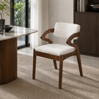 A modern wooden dining chair with curved armrests and white textured upholstery, placed in a bright, contemporary room next to a marble-topped table.