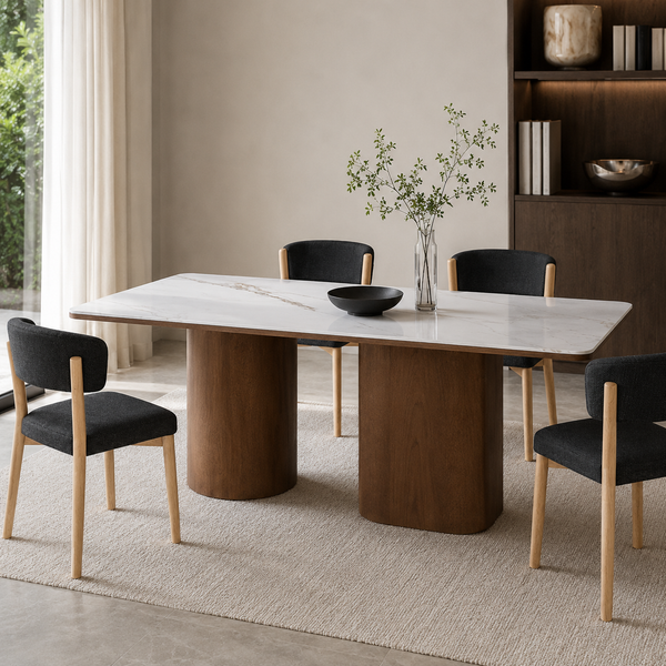 A modern dining table with a white marble-look top and thick cylindrical dark wood bases, surrounded by four black upholstered chairs with light wood legs in a bright, neutral-toned dining room.