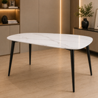 An oval dining table with a white marble-look top and black tapered legs, shown in a modern interior setting with wood flooring and warm ambient lighting.